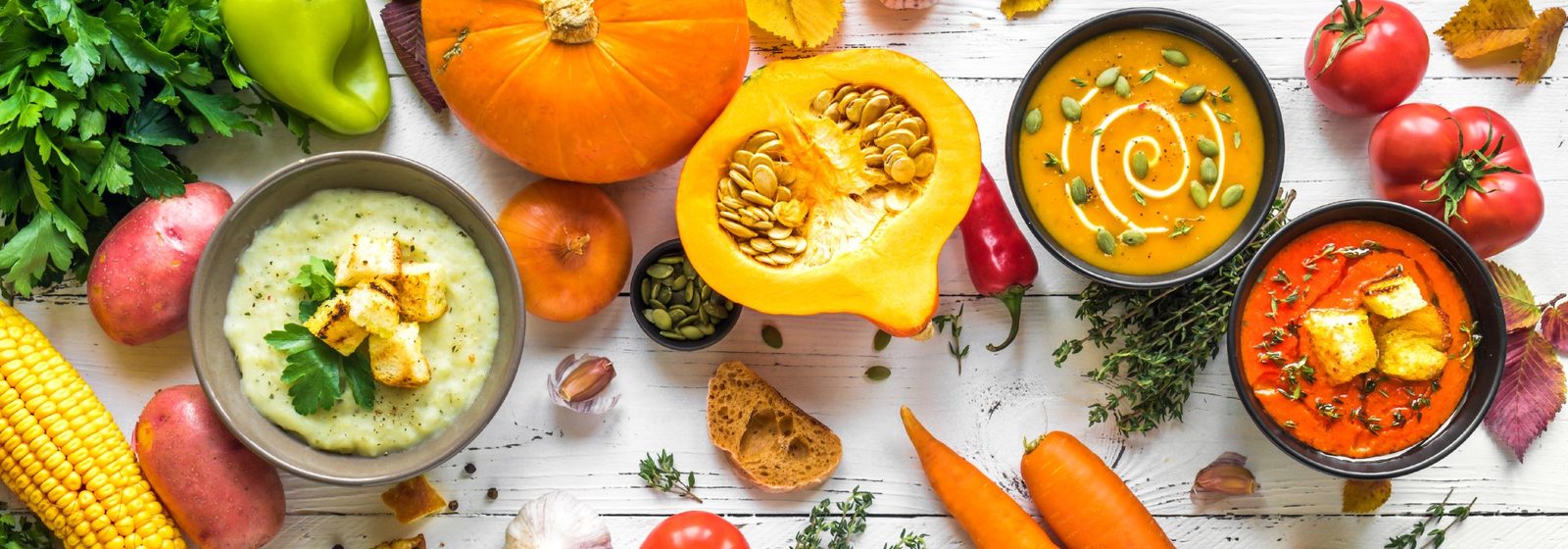 Herbstgemüse, Kürbis und drei Suppen in Schalen auf einem hellen Holztisch